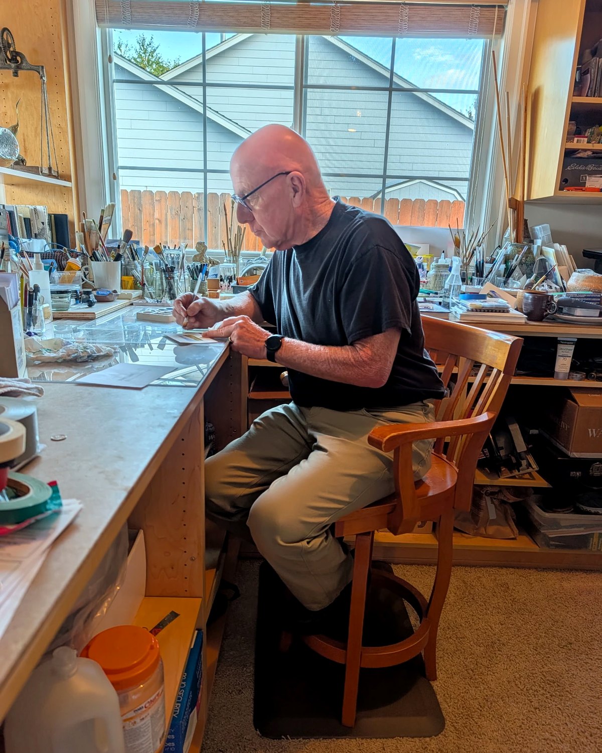 Richard Kuensting painting at his studio bench — brushes, palettes and the window light around him