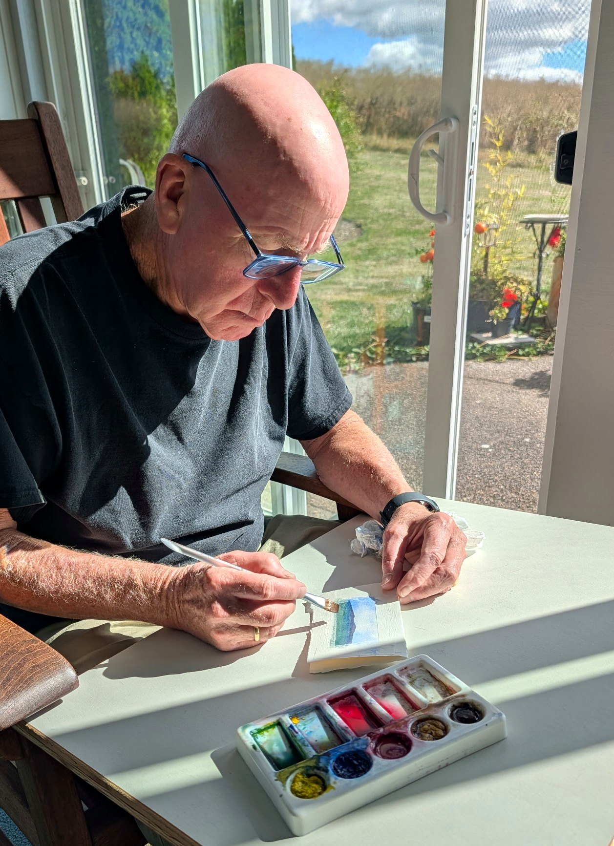 Richard painting at his bench by the studio window, the yard and fence beyond